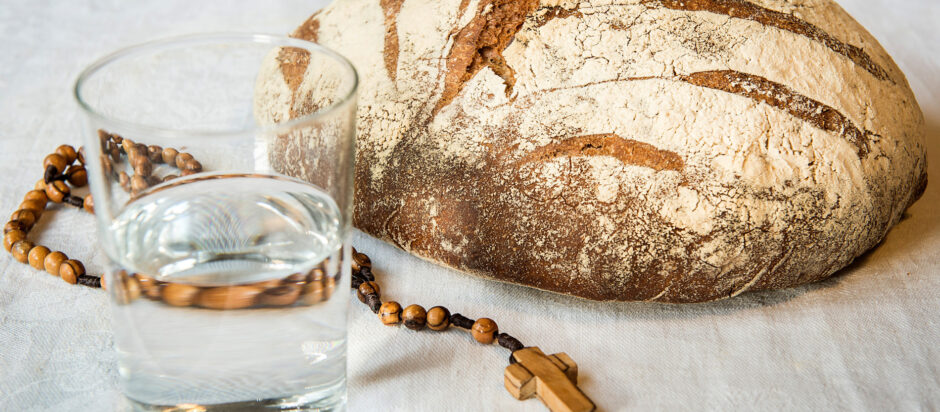 Ein Glas Wasser und ein Laib Brot. Dazwischen ein hölzerner Rosenkranz.